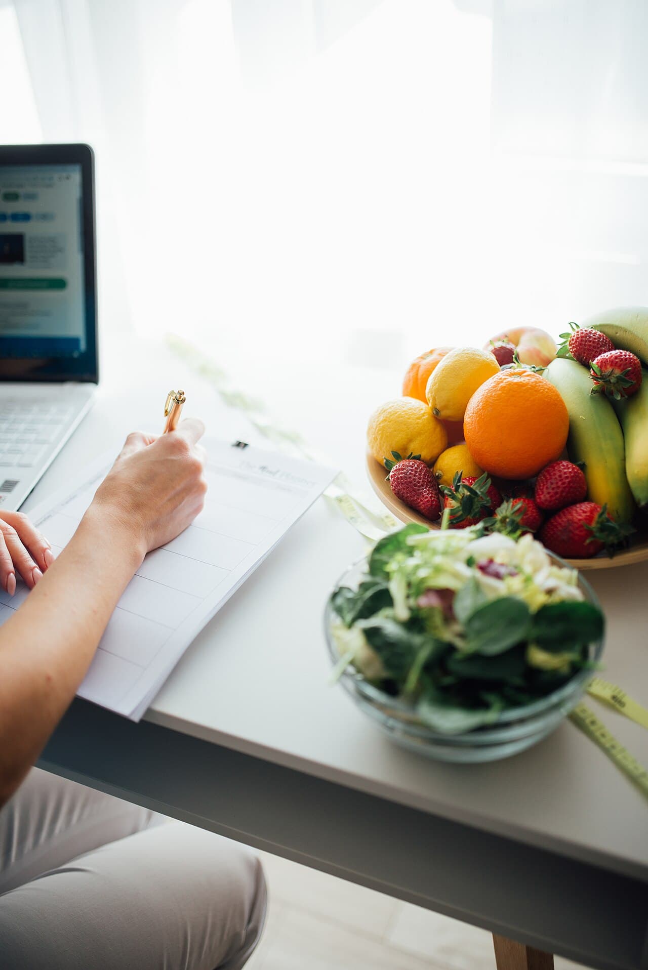 Pessoa anotando um planejamento de refeições ao lado de uma salada e de uma fruteira